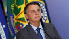 Bolsonaro com expressão séria falando ao microfone, com bandeiras no fundo