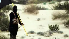 slamic State/Iraq/Syria: A masked Islamic State soldier poses holding the ISIL banner somewhere in the deserts of Iraq or Syria. ISIL publicity image, 2015. (Photo by: Pictures from History/Universal Images Group via Getty Images)