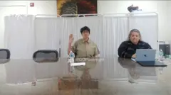 کیلین مکسول در جلسه استماعیه کنگره. او دست راست خود را بالا برده و لباس خاکستری به تن دارد. کنارش یک زن با لپ‌تاپ نشسته است. 