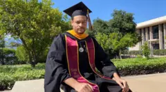 Cole Allen está sentado em um muro ao ar livre, sob o sol, em frente a uma área gramada com árvores. Ele olha para a câmera e está usando beca e capelo de formatura. A faixa vermelha e amarela diz "turma de 2025".