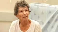 Yocheved Lifshitz sits in hospital bed holding cup, with two people standing either side of her