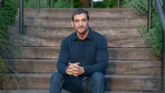 Amir Levine sentado em uma escada externa larga de madeira, levemente inclinado para frente com os dedos entrelaçados, vestindo jeans e uma camisa escura de mangas compridas