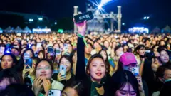 Fãs, em sua maioria mulheres, assistem à apresentação do Super Junior durante o Seoul Park Music Festival 2023. Uma fã segura o celular no alto e a tela exibe alguns caracteres coreanos.