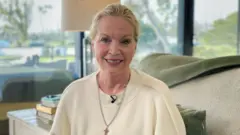 Una mujer rubia con una blusa color crema está sentada en un sofá sonriendo frente a las ventanas.