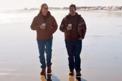 غريغ سكواير وبيت مانينغ يسيران على طول الشاطئ حاملين مشروبات جاهزة. كلاهما يرتديان بنطالاً أزرق وسترة بنية.