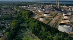 Vista aérea de uma grande refinaria de petróleo, com chaminés e tanques circulares de armazenamento, cercada por área de mata e um bairro residencial próximo
