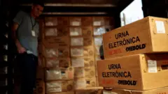 Pilhas de caixas de papelão com dizeres 'urna eletrônica'; um homem com crachá aparece no fundo da sala