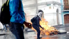 Dos personas protestan en Ecuador y de fondo se ve un colchón en llamas