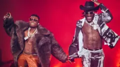 Wizkid and Burna boy performing on stage. Burna Boy is smiling and wearing a cowboy hat. Wizkid is wearing a big fur coat. Both have on sunglasses and jewellery. There is a red background.