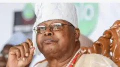 Aworan Oloogbe Ọba Sikiru Adetona Kayode, Awujale Ijebu. Oba naa jokoo lori itẹ pẹlu igo rẹ loju, ilekẹ oye lọrun, o si di awọn ika ọwọ rẹ pọ ninu aworan yii, o n wo orere.