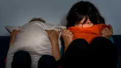 Duas mulheres vendo televisão sentadas na cama. Uma delas se cobre totalmente com uma almofada e a outra leva o cobertor à boca, mas continua assistindo atentamente com os olhos assustados