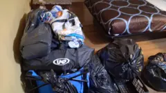 Bolsas de viagem e sacos de lixo cheios empilhadas no chão junto a uma cama com um edredon marrom