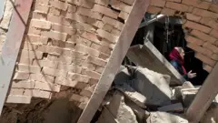 نمایی از یک ساختمان ویران شده در حملات آمریکا و اسرائیل به تهران و زنی با روسری قرمز که میان آواره‌ها ایستاده.