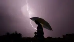 Pessoa caminha à noite com guarda-chuva ao ar livre com raio caindo ao fundo