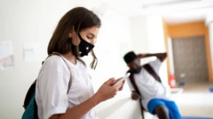 Em corredor de escola, menina mexe em celular, e menino aparece sentado atrás, ambos de uniforme