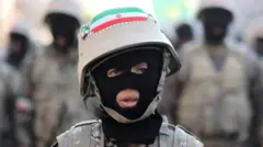 Un joven que lleva un pasamontañas, uniforme de fatiga y un casco con una bandera iraní mira fuera de cámara, con figuras militares borrosas en el fondo.