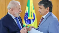 Lula conversa com o ministro do Desenvolvimento Social, Wellington Dias. Os dois têm expressão séria e Dias carrega um papel onde se lê "ações e programas". Ao fundo vê-se a bandeira do Brasil