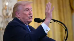 Trump, sarı bir arka planda konuşma yapıyor, önünde mikrofon var ve yalnızca kollarından üstü görebiliyoruz. Üzerinde takım elbise var. Tek eli havada.