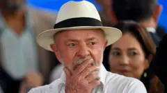 Lula, um homem idoso de barba grisalha, usa uma camisa branca e um chapéu branco de aba larga com faixa preta. Ele aparece em primeiro plano com a mão cobrindo parcialmente a boca e o queixo. Ao fundo, desfocado, há outras pessoas em um ambiente interno. A profundidade de campo reduzida mantém o foco no rosto e no gesto do homem.