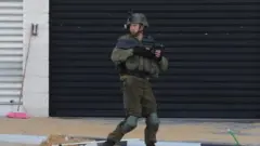 جندي إسرائيلي يمر من أمام المحلات المغلقة في الموقع حيث قتلت قواته ثلاثة فلسطينيين بالرصاص، بالقرب من حاجز صرة العسكري خارج مدينة نابلس بالضفة الغربية، 12 مارس/آذار 2023.