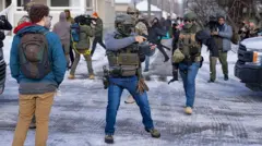 ضابط من هيئة الهجرة والجمارك الأمريكية (ICE) (على اليمين) وآخر من الضباط الفيدراليين يحمل جهازاً للتحكم في الحشود (في الوسط) يقفان عند تقاطع في مينيابوليس حيث تجمع المحتجون بعد مقتل رينيه نيكول غود برصاص واحد من عناصر الـICE، بينما يقف المارة والصحفيون داخل التقاطع كذلك.