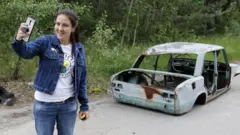 A visitor posses for a selfie next to a wrecked car at the Chernobyl exclusion zone