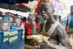 رجل يحدق في الكاميرا يرتدي القميص ويأكل أرزا خارج من حاوية بوليسترين