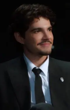 Jão, de cabelos escuros e bigode, veste terno preto, camisa branca e gravata preta, enquanto está sentado em uma poltrona. Ele sorri discretamente e olha para fora do enquadramento, em um ambiente escuro iluminado por luz direcionada. Copyright