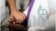 Pessoa com jaleco do Mais Médicos segura a mão de outra pessoa