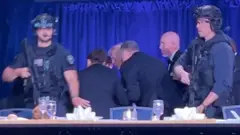 夕食会会場の主賓席の後ろで、トランプ氏がスーツ姿の男性たちに囲まれて身をかがめている。その周りをさらに、重装備の男性2人が自動小銃を構えて警備している