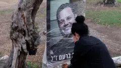 женщина в черном смотрит на портрет в мужчины в траурной рамке, который замотан прозрячной пленкой и прикреплен к дереву