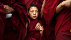 赤い僧服をまとったチベット仏教の子供の僧侶が、同じ格好の大人の僧侶たちに囲まれている。