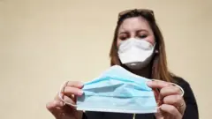 Una mujer con mascarilla en el rostro sosteniendo otra mascarilla