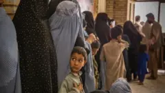 طفل يرافق أمه في طابور أمام منفذ توزيع للأطعمة تابع لأوكسفام