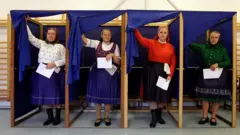 geleneksel Macar kıyafetleri giymiş kadınlar aynı anda oy kabininden çıkıyor. Hepsi renkli etekler ve bluzlar giymiş. Yaşları ileri görünüyor.