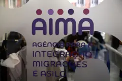 Logo da AIMA em uma agência, com pessoas sentadas aguardando atendimento em segundo plano