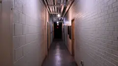 Longo corredor industrial, com paredes de blocos de concreto pintados de branco e piso de cimento, com canos percorrendo o teto
