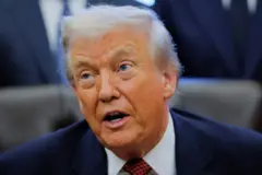 Близький портрет Трампа у темному діловому костюмі з білою сорочкою та червоно-чорною краваткою у клітинку. У тлі видно розмиті фігури інших людей у костюмах, що стоять позаду.