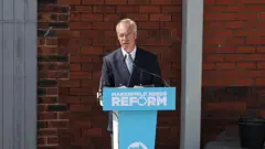El líder de Reform UK, Nigel Farage, habla en una conferencia de prensa sobre un atriz azul claro.