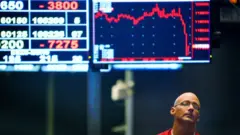 Borsaların çöküşünü gösteren kırmızı grafik ve rakamlardan oluşan ekranın önünde umutsuz bir yüz ifadesiyle duran gözlüklü ve kırmızı tişörtlü bir adam