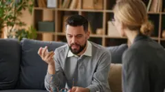 Homem sentado em um sofá com os óculos na mão fala a uma psicóloga durante uma sessão de terapia