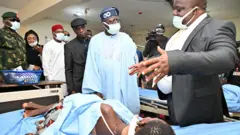 Nigeria President Bola Tinubu wear sky blue cloth and Navy blue cap, e astand opposite man wey wear back suit we be like say e dey explain something to am as e visit victims of di Benue killings for Hospital 