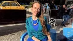 青系の服を着た長い髪の女性が屋外でいすに座りカメラを見てほほ笑んでいる。後背には多くの人や黄色い車が写っている