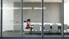 Uma mulher de vestido vermelho está sentada em um escritório com cadeiras brancas, trabalhando em seu laptop