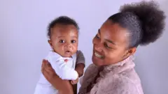 Mãe sorri enquanto olha e segura seu bebê recém-nascido, que olha com cara de assustado para a câmera
