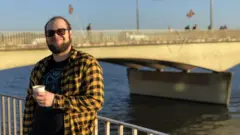 cosmólogo brasileiro Pedro Bernardinelli em frente a uma ponte nos Estados Unidos, tomando café. Está vestido com uma camisa xadres (amarela e preta) e camiseta vermelha por baixo; usa óculos escuros.