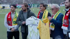 Dirigentes do clube, com cachecol que mistura as cores do Exeter e do Brasil, trocam camisas em dia de celebração da história do clube que enfrentou a seleção em 1914