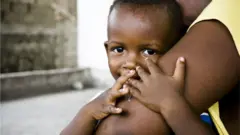 Bebê segurando o braço de sua mãe e olhando para a câmera
