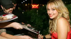 Hayden Panettiere no tapete vermelho da festa Teen Vogue Young Hollywood, distribuindo autógrafos. Ela está usando um vestido vermelho e sorrindo para a câmera.