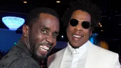 Close up view of Sean 'Diddy' Combs and Jay-Z smiling at a music industry event. They are in a dark interior and are both wearing suits. Jay Z is wearing sunglasses